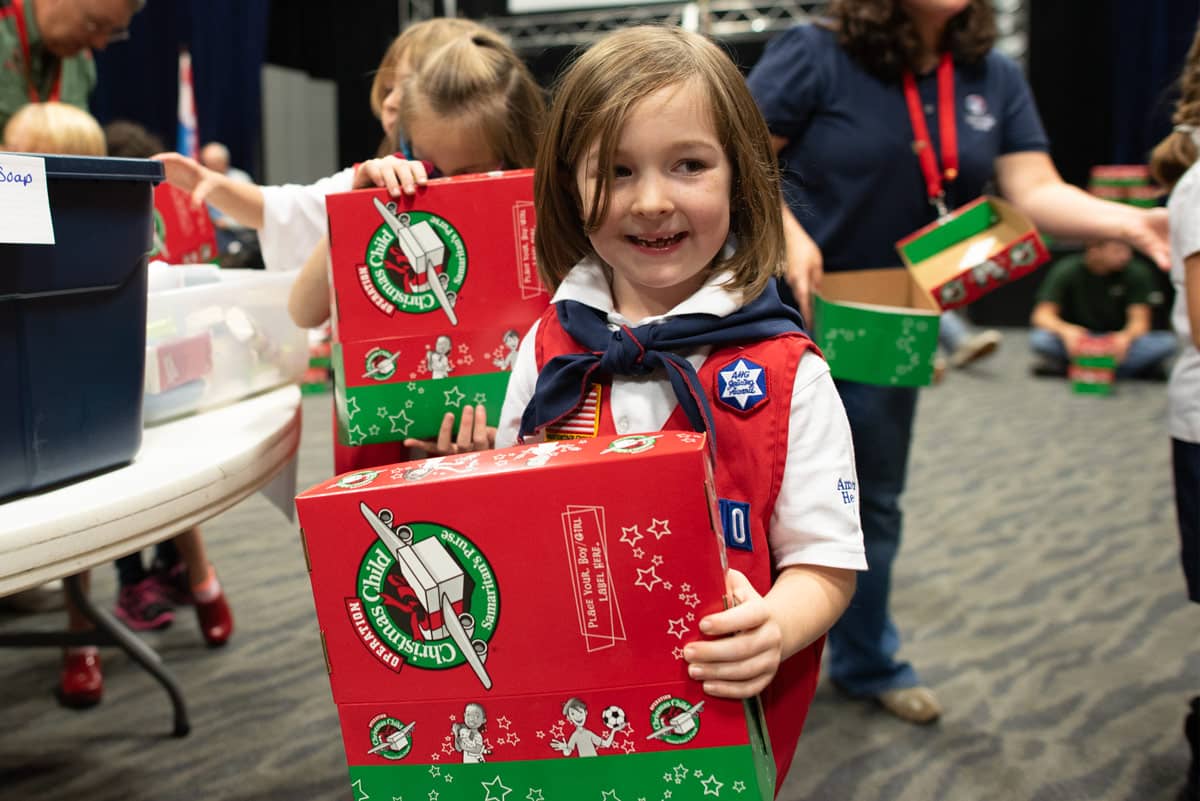 Little girl in an American Heritage Girls uniform holding a shoebox and smiling.