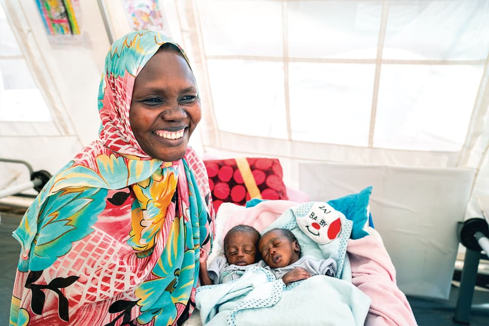 A mother smiling with two babies sleeping in a crib.