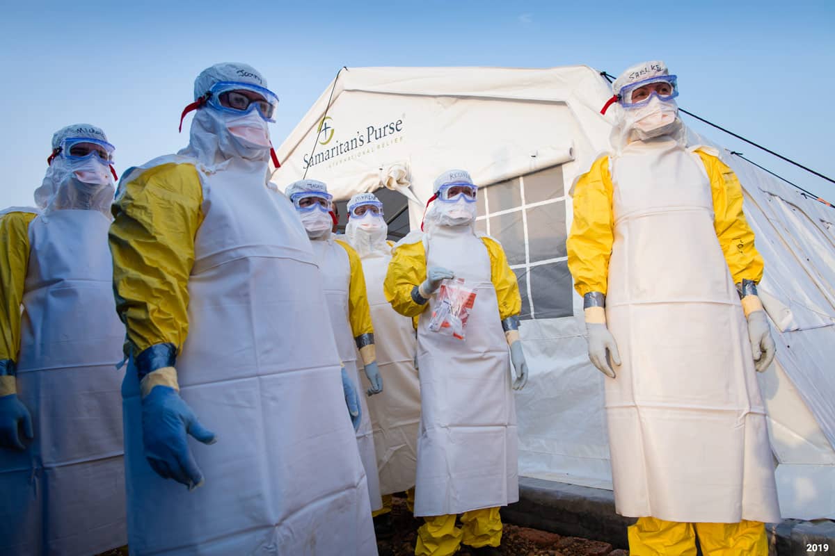 A group of medical workers in full body sanitation suites standing in front of a Samaritan's Purse medical tent.
