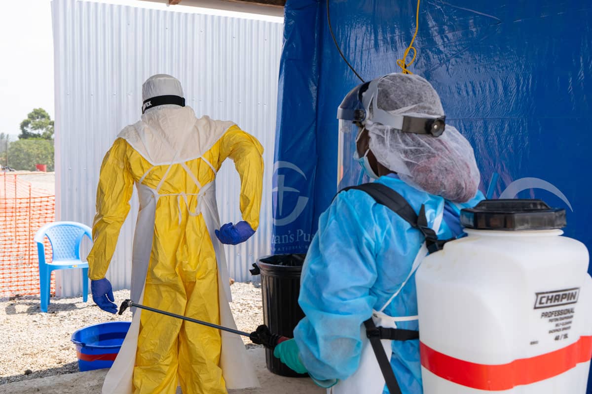 One team member sprays down medical personnel who has exited the patient area. This bleach solution is part of the decontamination protocol before removing protective gear.