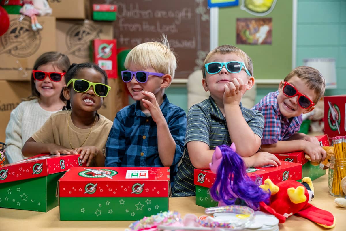 Two girls and three boys making faces with sunglasses in front of shoeboxes.