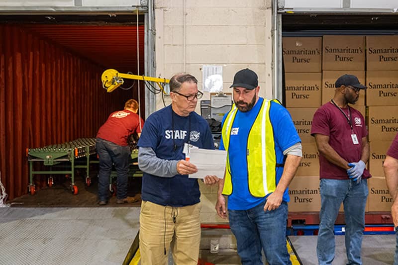 Two men discusing and holding a paper with the plans for the shipment of the large tan shipping boxes.