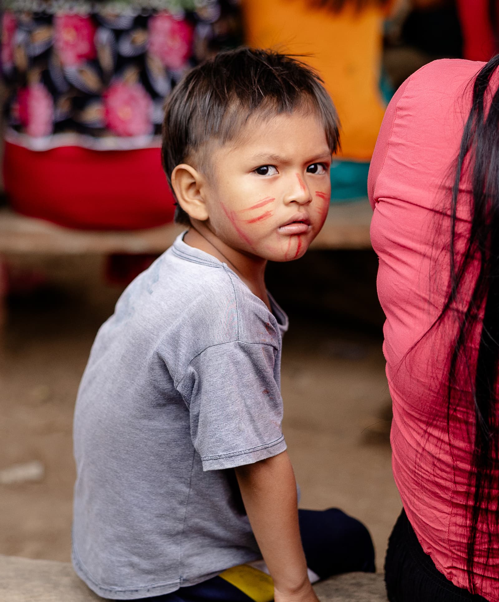 A little boy with red face paint markings frowning while sitting on a bench.