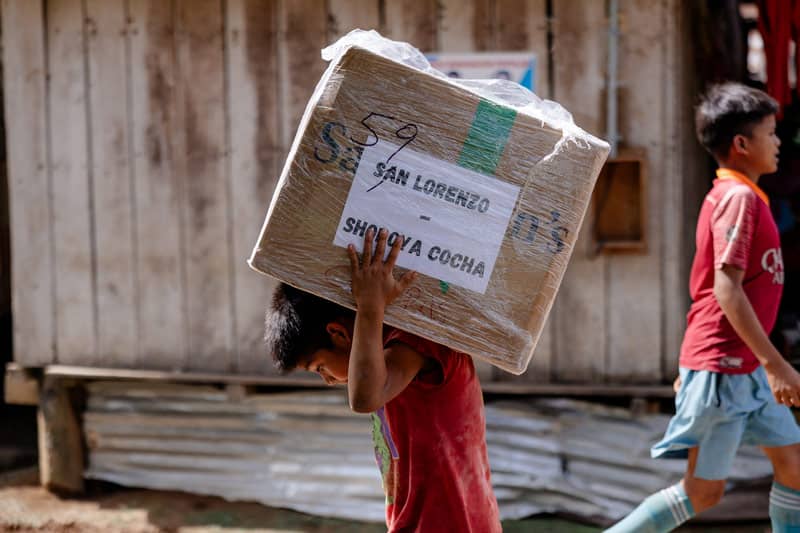 A boy carrying a large tan box into village.