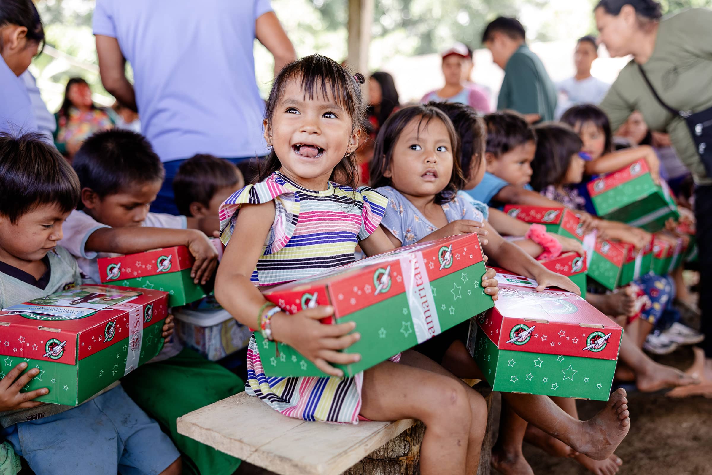 A little girl smiling while opening her shoebox gift with a large group of children behind her.