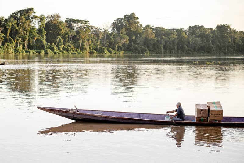 A man sitting in a canoe with multiple large tan boxes in the back of the canoe.