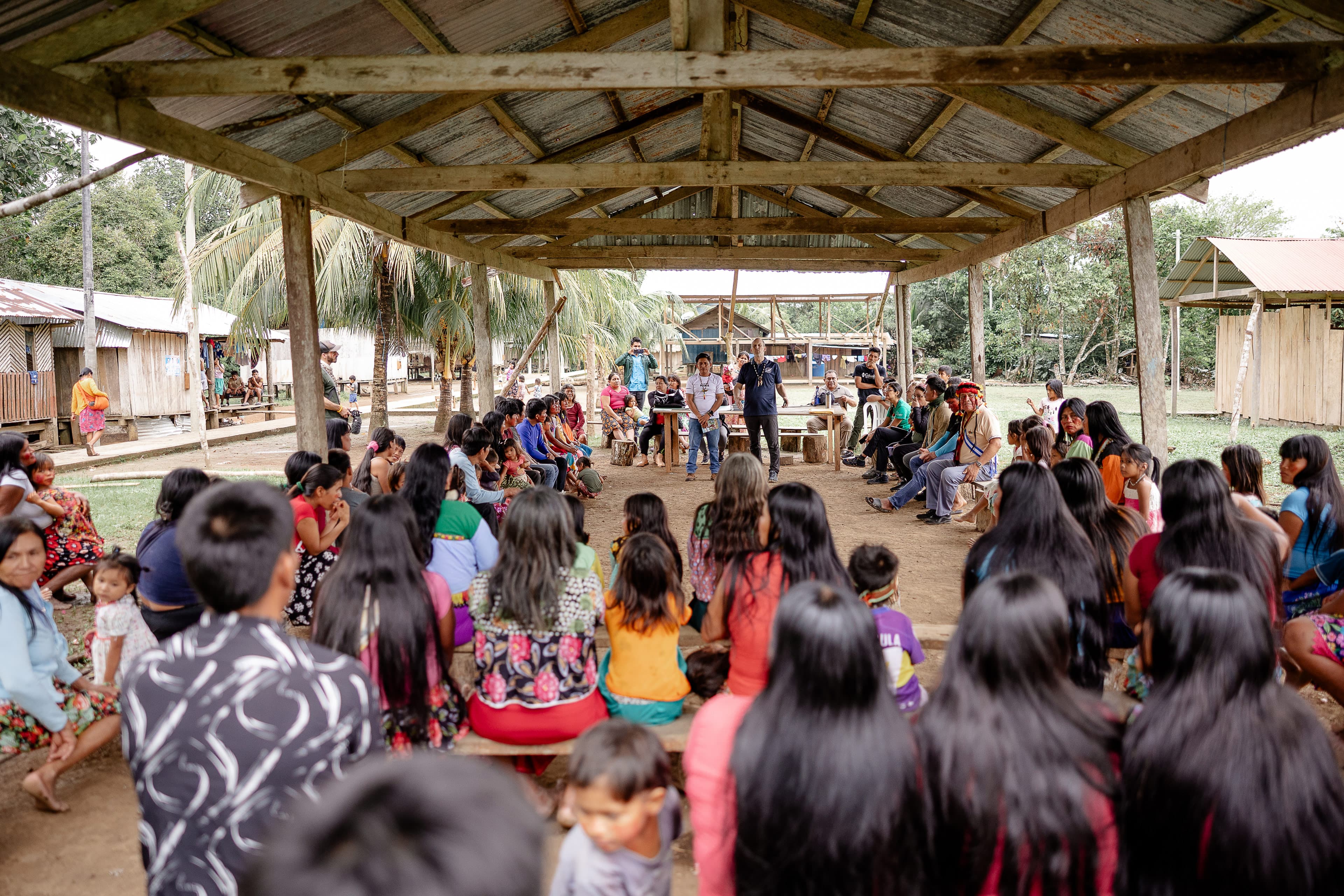 Two men preaching the gospel to a group of people under a covered roof.
