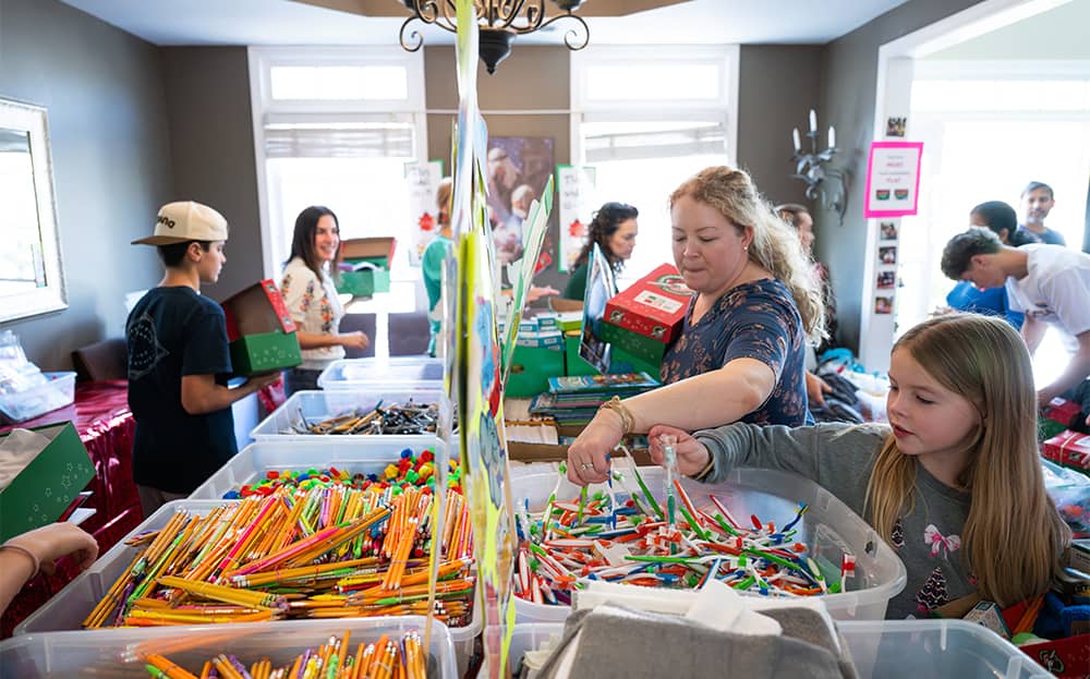 Adults and children packing shoeboxes with gifts from bins on a table in a house.
