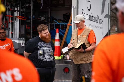 Volunteer in orange shirt and tan vest reading a bible to homeowner.
