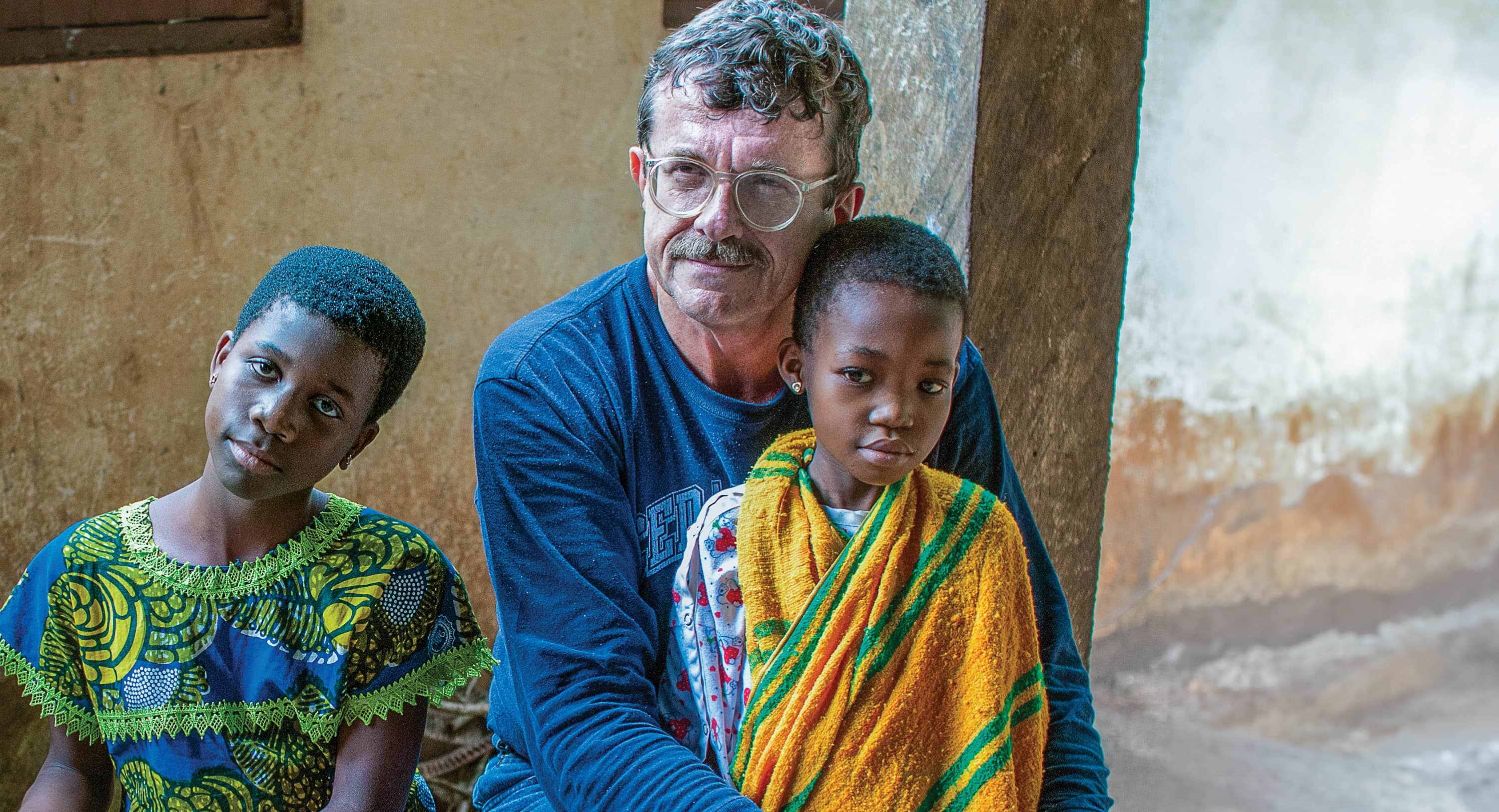 A man with glasses seated alongside two young children inside a rustic building.