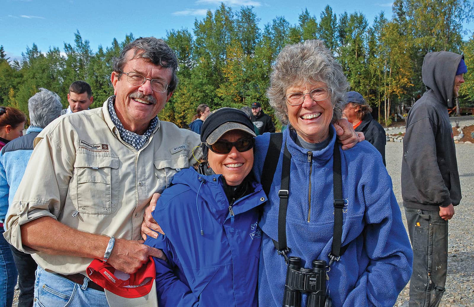Three smiling people pose for a photograph outdoors on a sunny day, with bystanders in the background.